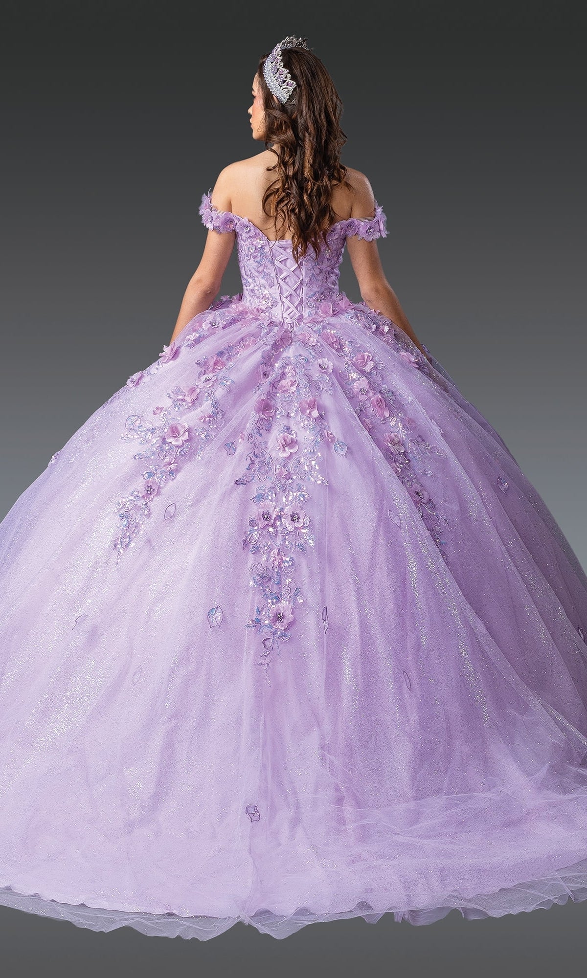 A woman in the Quinceanera Dress 1951 By Dancing Queen, a sparkly lilac purple off-the-shoulder ball gown with floral appliquรฉs and a tiara, stands facing away to showcase the skirt against a gray background.